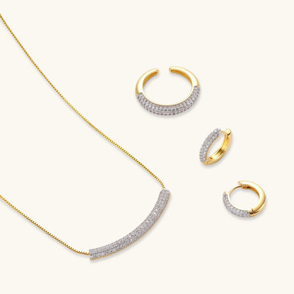 Gold-plated curved necklace, hoop earrings, and adjustable ring with pavé cubic zirconia stones on cream background.