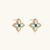 Gold quatrefoil stud earrings with white mother-of-pearl petals and emerald green gemstone center on beige background.