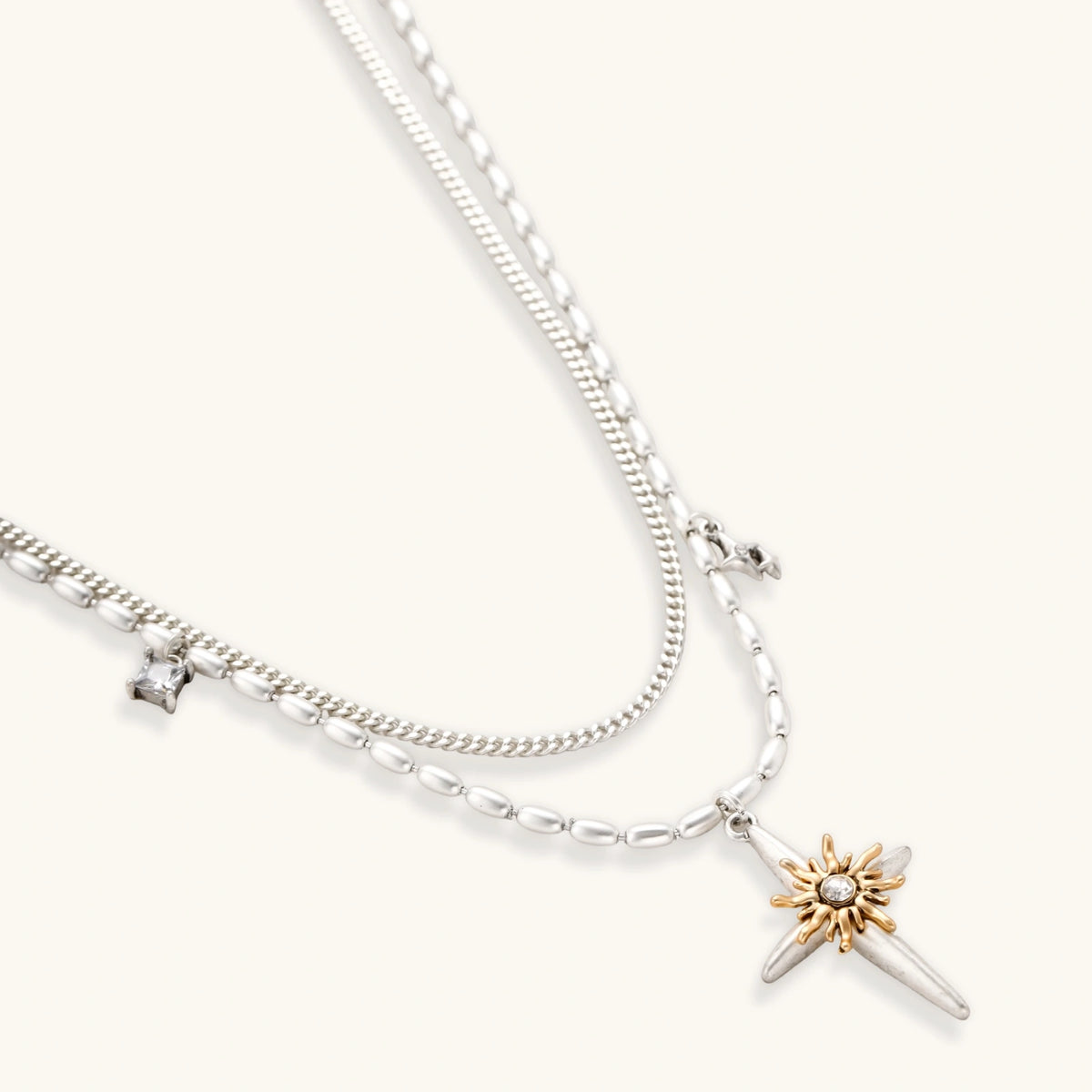 Layered silver-toned necklace set with cross pendant and gold sun charm detail, matching drop earrings on white background.
