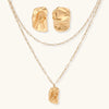 Sculptural gold-toned jewelry set with organic fluid shapes including earrings and layered chain necklace on cream background.