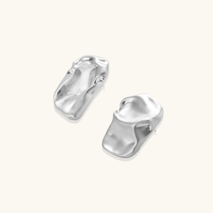 Pair of sculptural silver ear cuffs with organic fluid shapes and polished finish against cream background.