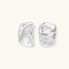 Pair of sculptural silver ear cuffs with organic fluid shapes and polished finish against cream background.