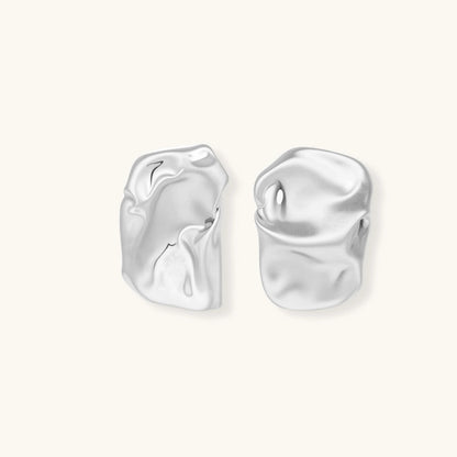 Pair of sculptural silver ear cuffs with organic fluid shapes and polished finish against cream background.