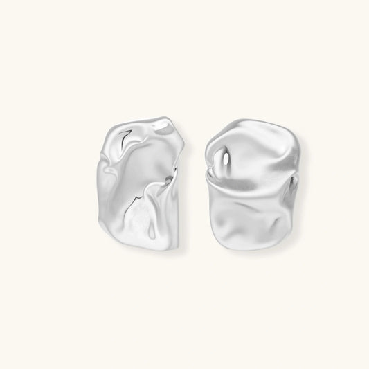 Pair of sculptural silver ear cuffs with organic fluid shapes and polished finish against cream background.
